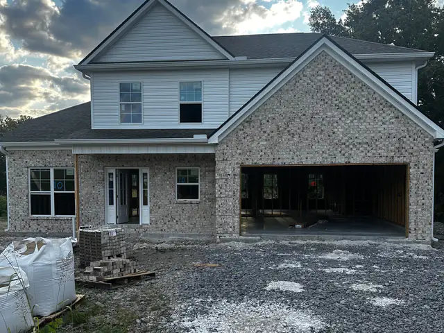 Two-story home under construction with light brick, white siding, and gravel driveway